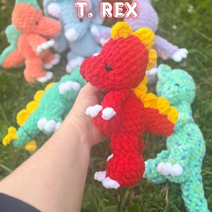 May include: A collection of crocheted dinosaur plush toys in various colors including red, orange, blue, green, and purple. The central red T. REX figure features yellow dorsal spikes and white claws. The text Remi T. REX is visible above the toys.