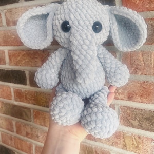 May include: A soft, gray crocheted elephant plush toy with black button eyes. The elephant is sitting in a person's hand.