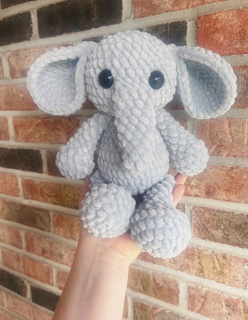 May include: A soft, gray crocheted elephant plush toy with black button eyes. The elephant is sitting in a person's hand.