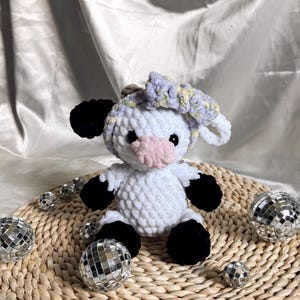 May include: White crocheted cow plush toy with black hooves and ears. The cow is wearing a purple and white crocheted flower headband. The toy is sitting on a woven straw mat with silver disco balls.