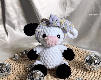 May include: White crocheted cow plush toy with black hooves and ears. The cow is wearing a purple and white crocheted flower headband. The toy is sitting on a woven straw mat with silver disco balls.