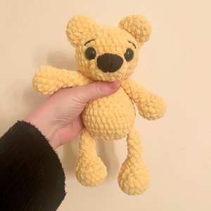 May include: A crocheted yellow teddy bear with black eyes and a black nose. The bear has a fluffy texture and is standing on its four legs.