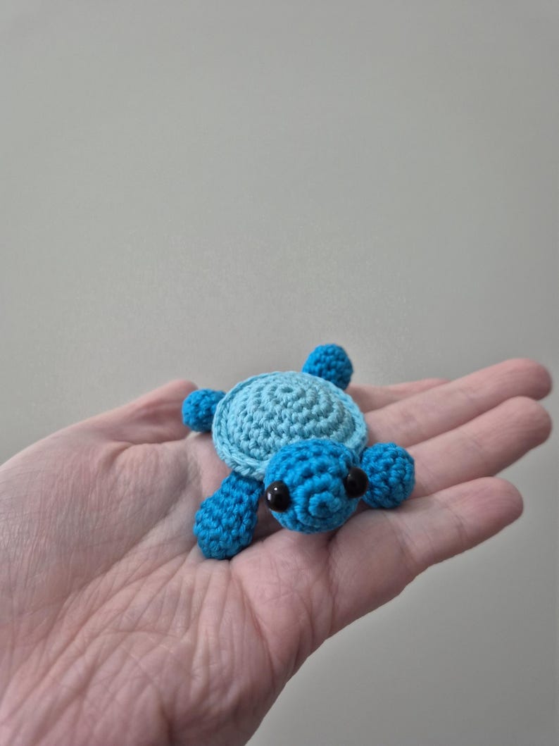 May include: A small, handmade crochet turtle in shades of blue. The turtle has a light blue shell, dark blue flippers and head, and black eyes. The turtle is a cute, whimsical design.