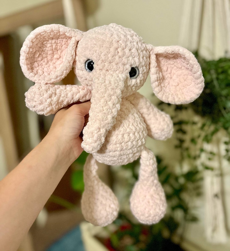 May include: A pink crocheted elephant plush toy with black eyes. The elephant has a long trunk and floppy ears.