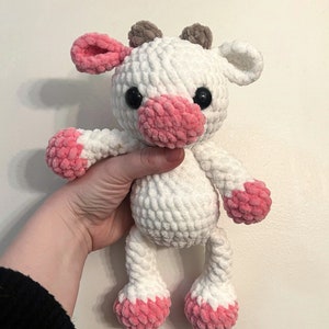 May include: White crocheted plush toy cow with pink accents on the ears, nose, and feet. The cow has black eyes and brown horns.