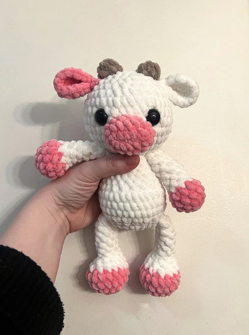 May include: White crocheted plush toy cow with pink accents on the ears, nose, and feet. The cow has black eyes and brown horns.