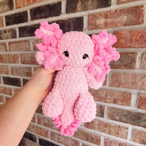 Puede incluir: Un peluche de axolote tejido a ganchillo en color rosa claro con un acabado de hilo suave y texturizado. La criatura presenta ojos grandes, oscuros y brillantes, y prominentes branquias externas rizadas a ambos lados de la cabeza en rosa oscuro.