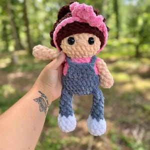 May include: A crocheted doll with brown hair, a pink headband, a pink shirt, blue overalls, and white shoes.