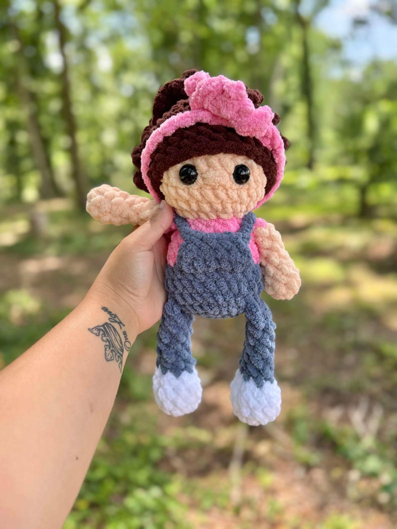 May include: A crocheted doll with brown hair, a pink headband, a pink shirt, blue overalls, and white shoes.