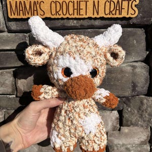 May include: A crocheted brown and white cow plush toy with black eyes. The toy is standing on a stone surface. The text "MAMA'S CROCHET N CRAFTS" is visible in the background.