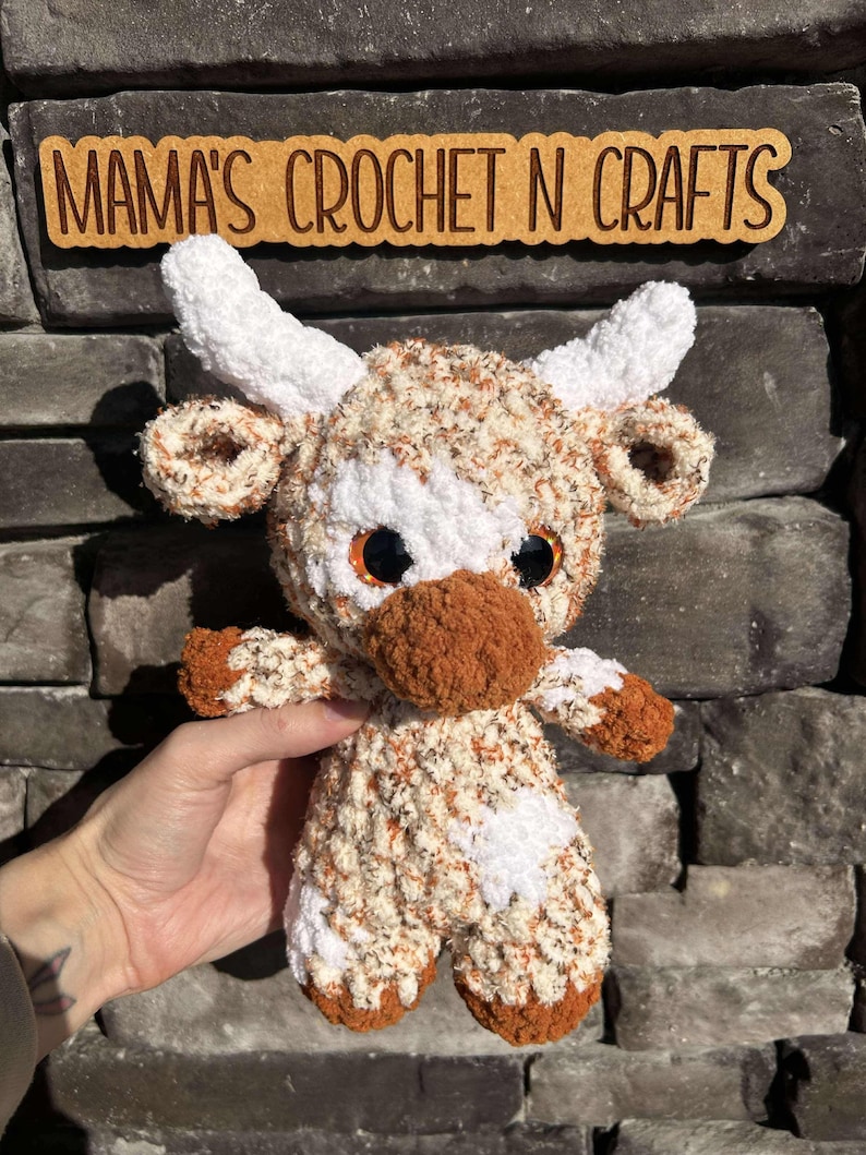 May include: A crocheted brown and white cow plush toy with black eyes. The toy is standing on a stone surface. The text "MAMA'S CROCHET N CRAFTS" is visible in the background.