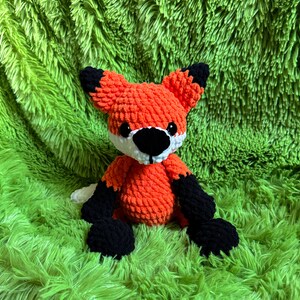 May include: A crocheted orange fox plush toy with black and white details. The toy is sitting on a green fuzzy surface.