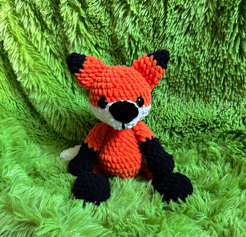 May include: A crocheted orange fox plush toy with black and white details. The toy is sitting on a green fuzzy surface.