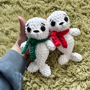 Puede incluir: Dos peluches de foca de crochet blanco con ojos negros. Una foca lleva una bufanda verde y la otra una bufanda roja.