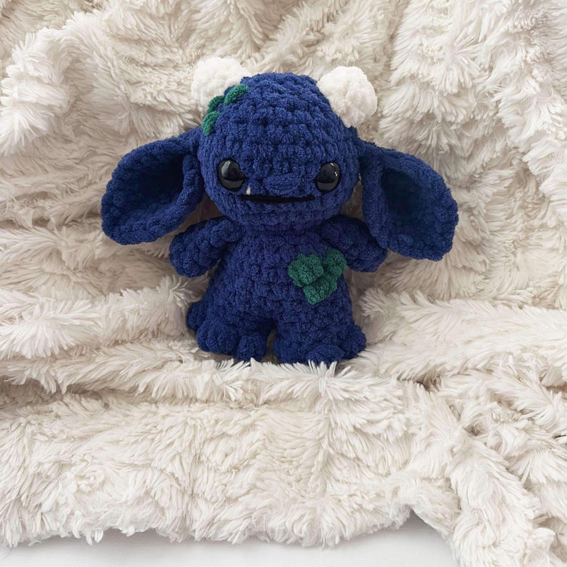 May include: A blue crocheted plush toy with white horns and green leaves. The toy has large black eyes and a toothy grin. It is wearing a green heart on its chest.