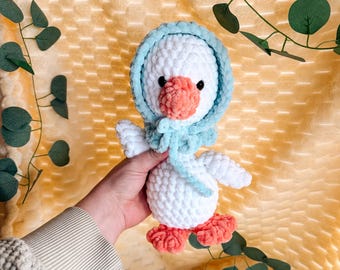 May include: A handmade white crochet goose wearing a light blue bonnet and scarf. The goose has a peach-colored beak and feet. The plush toy is soft and fluffy, with a detailed design. The background is a textured yellow surface with green leaves.