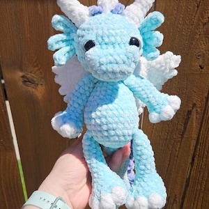 May include: A crocheted blue dragon with white wings and purple accents. The dragon has large black eyes and a fluffy texture.