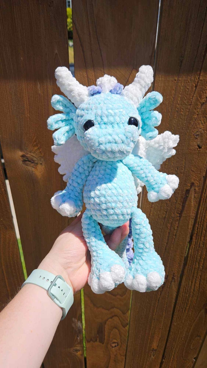 May include: A crocheted blue dragon with white wings and purple accents. The dragon has large black eyes and a fluffy texture.