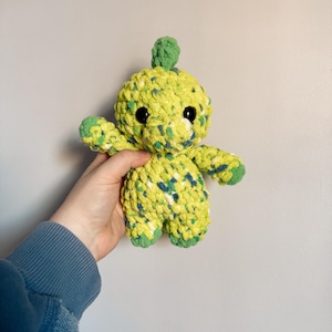 May include: A green and yellow crocheted dinosaur plush toy with black eyes. The toy is being held in a hand.