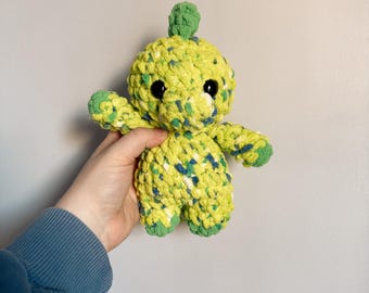 May include: A green and yellow crocheted dinosaur plush toy with black eyes. The toy is being held in a hand.