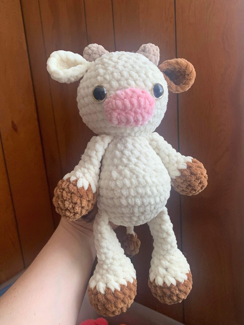 May include: A white crocheted cow plush toy with brown ears and hooves. The cow has pink felt nose and black button eyes.