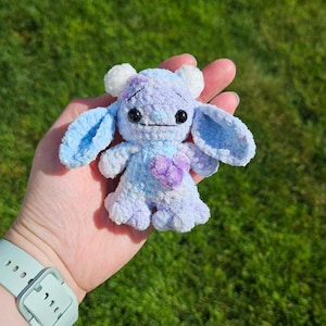 May include: A small, crocheted plush toy in the shape of a monster. The toy is light blue and purple with white horns and a purple heart on its chest.