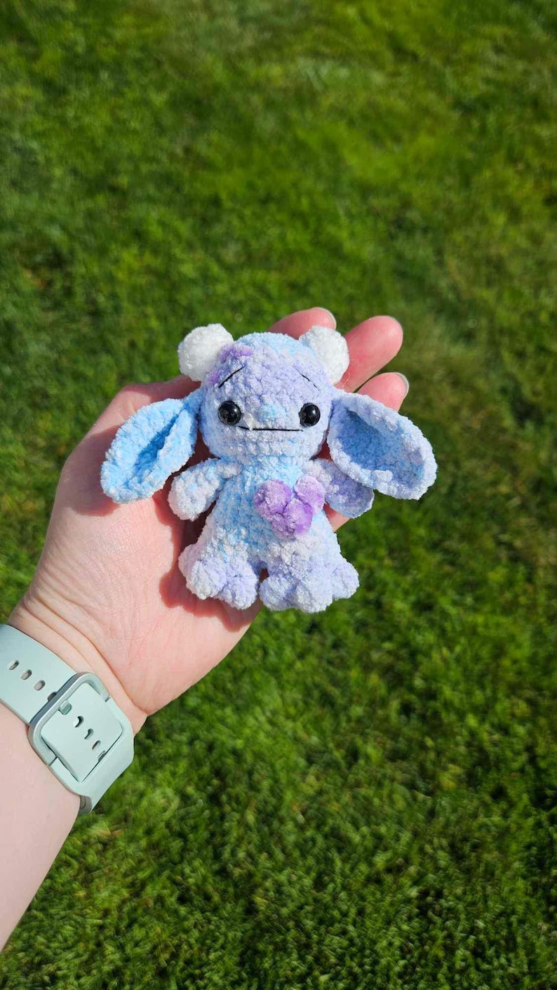 May include: A small, crocheted plush toy in the shape of a monster. The toy is light blue and purple with white horns and a purple heart on its chest.
