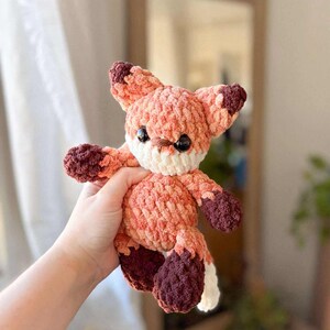 May include: A crocheted fox toy in orange, peach, and brown. The fox has a white muzzle and tail tip, with dark brown accents on the ears and paws. The toy is soft and cuddly, suitable for a child's room or as a decorative item. The fox is handmade.