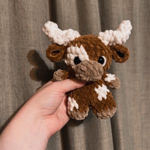 May include: A brown and white crocheted stuffed animal shaped like a cow with large white horns. The cow has black eyes and a brown nose.