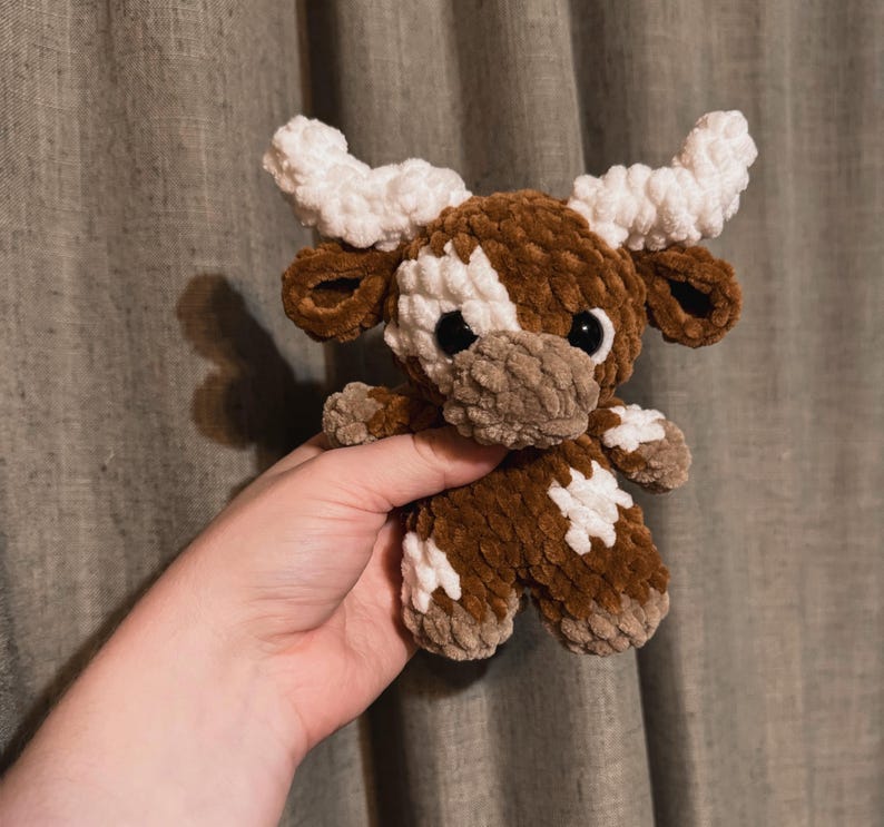 May include: A brown and white crocheted stuffed animal shaped like a cow with large white horns. The cow has black eyes and a brown nose.