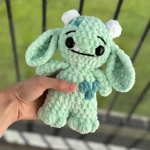 May include: A green crocheted plush toy with a friendly expression. The toy has large eyes, a wide smile, and two small horns on its head. It is holding a small blue heart in its hands.