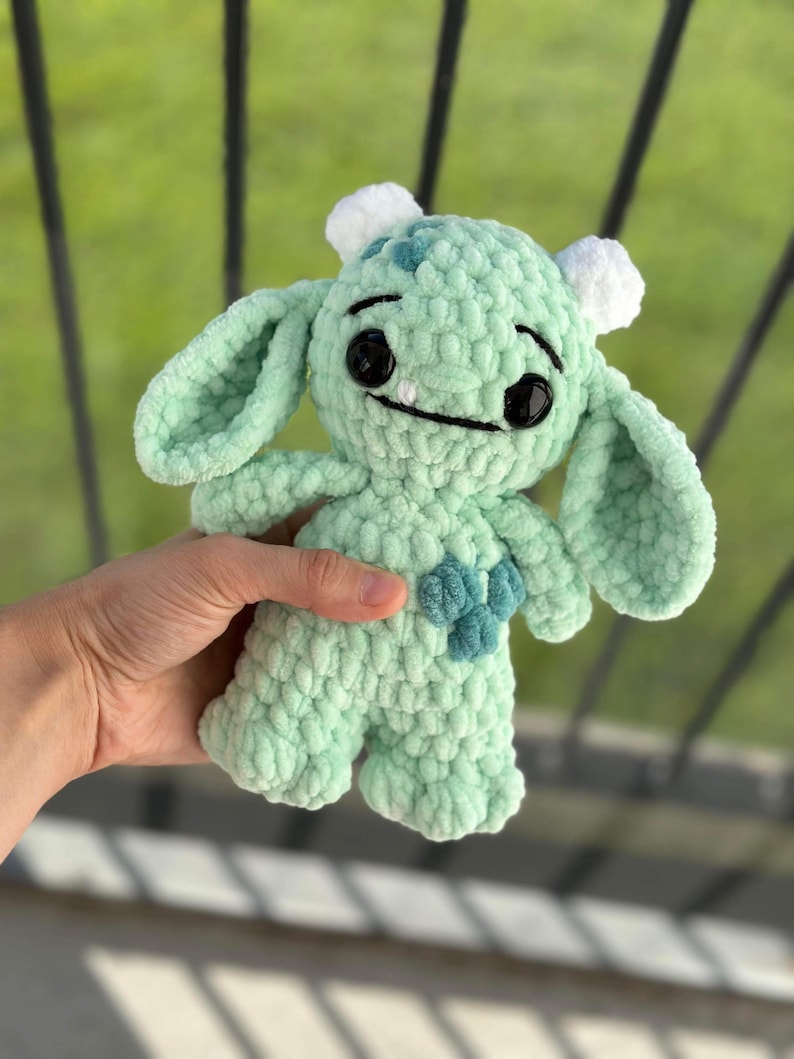 May include: A green crocheted plush toy with a friendly expression. The toy has large eyes, a wide smile, and two small horns on its head. It is holding a small blue heart in its hands.