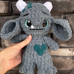 May include: A gray crocheted plush toy with large blue eyes and a green heart on its chest. The toy has white crocheted horns and ears. The toy is sitting on a brick wall with a sign that says "Mama's Crochet N Crafts".