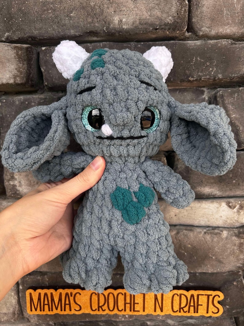 May include: A gray crocheted plush toy with large blue eyes and a green heart on its chest. The toy has white crocheted horns and ears. The toy is sitting on a brick wall with a sign that says "Mama's Crochet N Crafts".