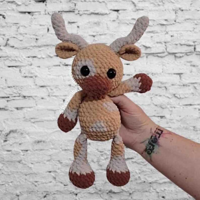 May include: A handmade crochet deer plushie in shades of tan, brown, and white. The deer has large black eyes, a brown nose, and small antlers. The plushie is being held up by a hand.