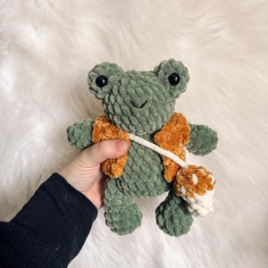 May include: A green crocheted frog plush toy wearing a brown crocheted vest and holding a white and brown crocheted pom-pom.