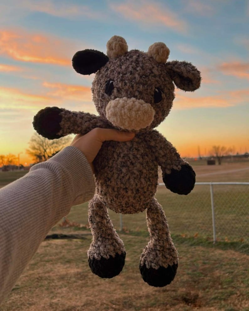 May include: A brown and black plush cow toy with a white nose and black eyes. The toy is being held up in the air against a sunset sky.