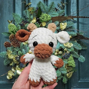 May include: A white crocheted stuffed animal shaped like a cow with brown accents. The cow has large black eyes and a brown nose. The cow is being held in a hand against a background of a green Christmas wreath.