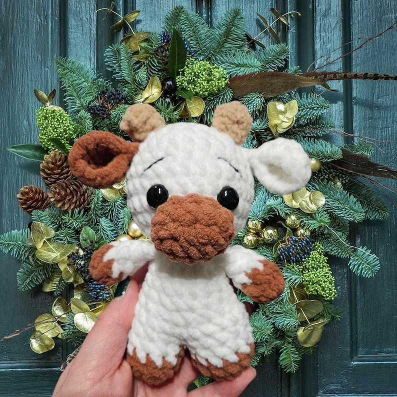 May include: A white crocheted stuffed animal shaped like a cow with brown accents. The cow has large black eyes and a brown nose. The cow is being held in a hand against a background of a green Christmas wreath.