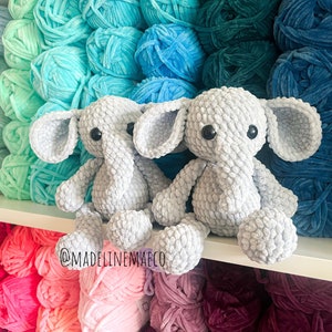 May include: Two gray crocheted elephant plushies sitting on a shelf with colorful yarn balls in the background. The yarn balls are arranged in a rainbow pattern. The plushies have black eyes and are made with a textured yarn. The text "@madelinenmaeco" is visible on the shelf.