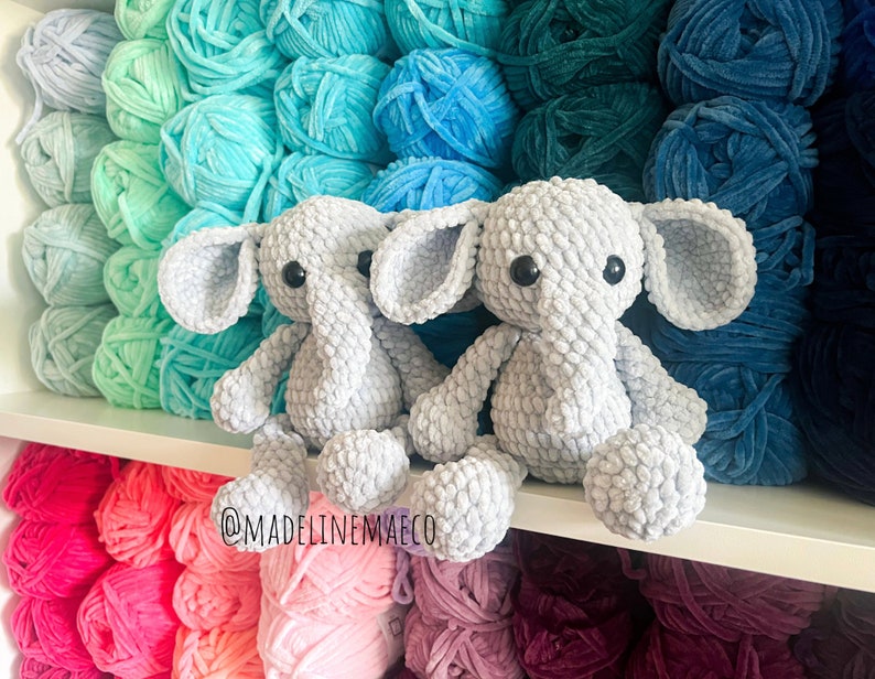 May include: Two gray crocheted elephant plushies sitting on a shelf with colorful yarn balls in the background. The yarn balls are arranged in a rainbow pattern. The plushies have black eyes and are made with a textured yarn. The text "@madelinenmaeco" is visible on the shelf.