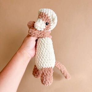Puede incluir: Un peluche de hurón hecho a mano con ganchillo en tonos crema, marrón y beige. El hurón tiene una cabeza crema con detalles marrones, un cuerpo crema y marrón, y una cola beige. El peluche está sostenido por una persona.