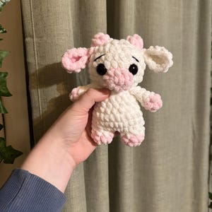 以下が含まれることがあります： ピンクの耳と鼻を持つ、白の毛糸で編まれたぬいぐるみ牛。牛は黒い目と小さな白い体を持っています。