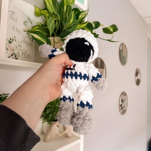 May include: A handmade white and blue crochet astronaut doll with a black visor. The doll has gray accents on the hands and feet. The astronaut is being held in front of a shelf with a plant and a sign that says "This is our Happy Place."
