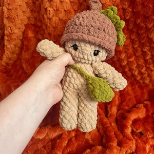 Puede incluir: Una muñeca de ganchillo hecha de hilo color canela suave, con un sombrero marrón en forma de bellota con una pequeña hoja verde en la parte superior. La muñeca tiene ojos de seguridad negros y lleva un pequeño bolso cruzado verde texturizado.