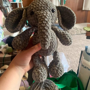 May include: A gray crocheted elephant plush toy with black eyes. The elephant is being held by a hand. The toy is sitting on top of a ball of gray yarn.