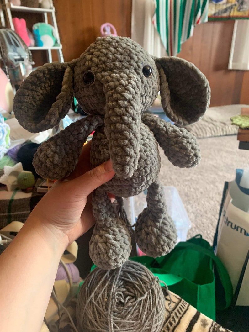 May include: A gray crocheted elephant plush toy with black eyes. The elephant is being held by a hand. The toy is sitting on top of a ball of gray yarn.