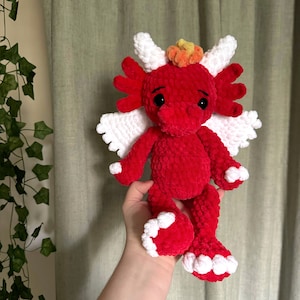 Könnte beinhalten: Ein roter gehäkelter Drache mit weißen Flügeln und einer gelben und orangenen Krone. Der Drache hat schwarze Augen und eine kleine, rosafarbene Nase.