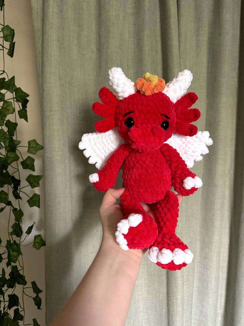 May include: A red crocheted dragon with white wings and a yellow and orange crown. The dragon has black eyes and a small, pink nose.