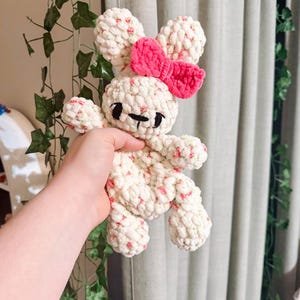 May include: A handmade crochet bunny plushie. The bunny is cream-colored with red and green speckles, a pink bow, and black embroidered facial features. The plushie is held in a hand.